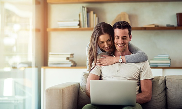 Are you expecting a tax refund? Make it count! Shot of a young woman hugging her husband while he uses a laptop on the sofa at home.