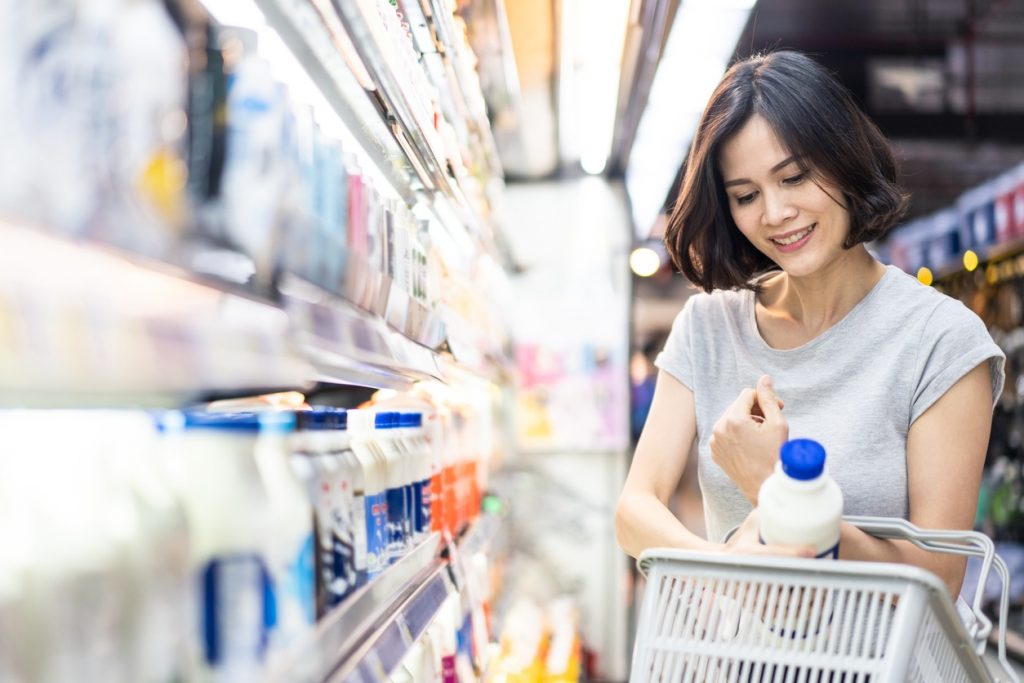 The Grocery Store Can Have a Big Impact on Your Budget | Young Asian beautiful woman holding grocery basket walking in supermarket. She is choosing daily milk product picking up from shelf. Seen from side while she looking at products. Shopping concept.